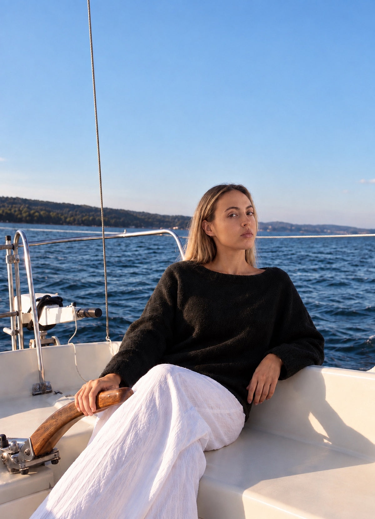 Merry Boatneck Sweater in Black, seated on a sailboat with white trousers, closer view.