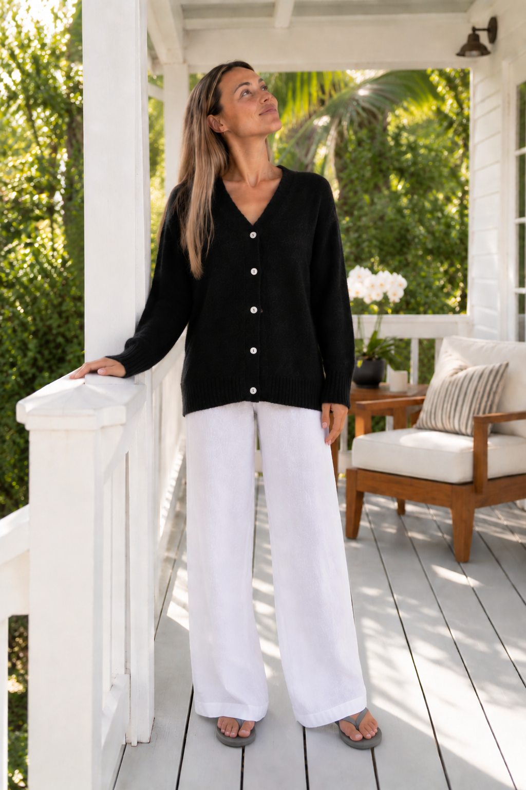 Frost Cardigan in Black, standing on a porch with white trousers.