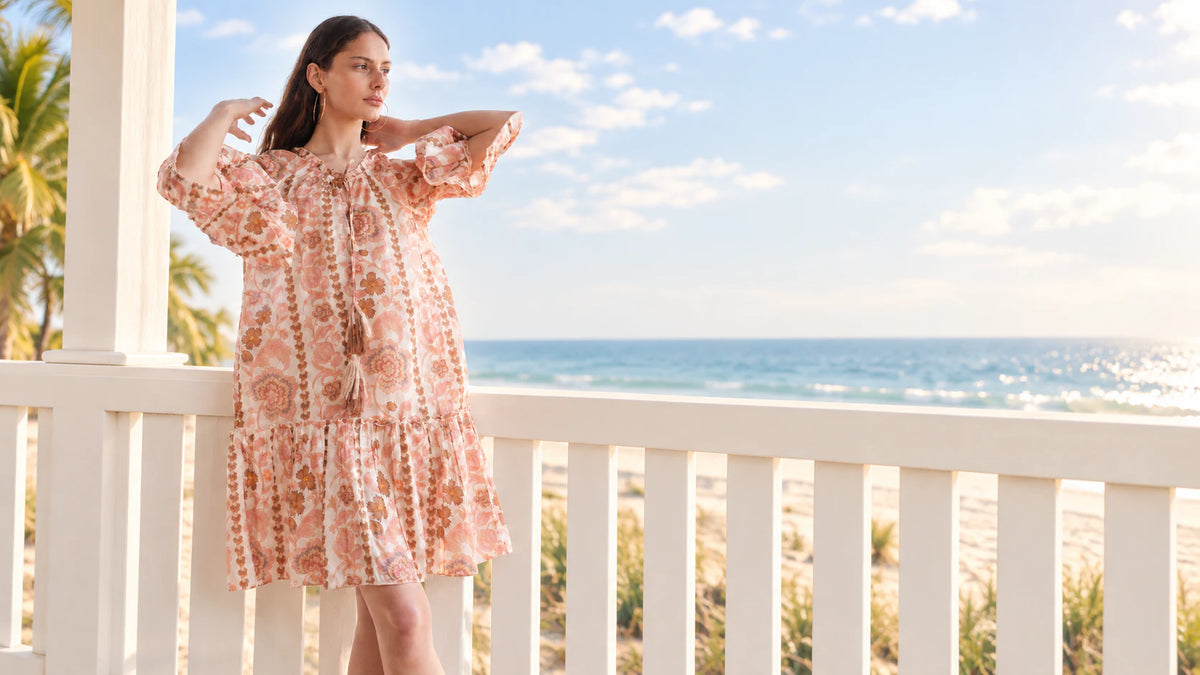 Woman wearing a peach floral resort dress on a sunlit balcony by the ocean
